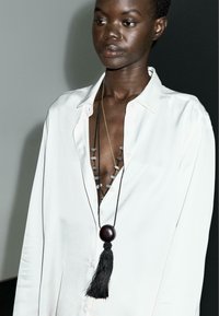 Camicia bianca lunga con scollatura a V profonda, adornata con elementi decorativi grigi e collane sovrapposte. Una collana presenta una grande perlina scura e un tassello nero.