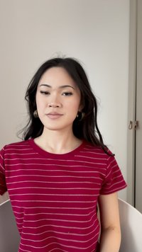 Junge Frau mit langen dunklen Haaren, die ein rot-weiß gestreiftes Shirt und Creolen trägt, sitzt vor einem schlichten hellen Wandhintergrund.