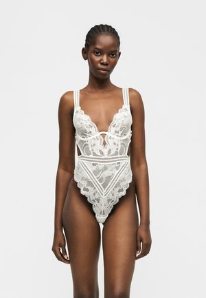 Γυναίκα που φοράει λευκό κορμάκι δαντέλας με λουλουδάτα σχέδια, ράντες στους ώμους και ψηλές ανοίγματα στα πόδια, στέκεται μπροστά σε απλό φόντο.