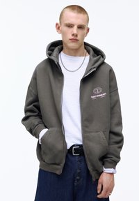 Fiatal férfi rövid hajjal, szürke, nyitott cipzáras kapucnis pulóvert visel fehér póló fölött, sötét farmernadrágot gombos sliccel, és ezüst színű láncnyakláncot.