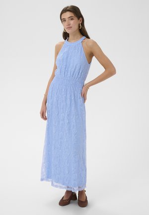 Mladá žena v priliehavých svetlomodrých čipkovaných maxi šatách bez rukávov a hnedých tkaných topánkach pózuje s rukou na bedre na bielom pozadí.