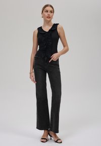 Schwarzes ärmelloses Oberteil mit Rüschenfront, kombiniert mit dunklen gerade geschnittenen Jeans und schwarzen offenen Schuhen. Weißer Hintergrund.