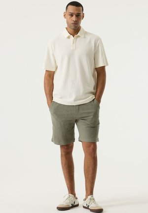 Crèmekleurig poloshirt met korte mouwen, gemaakt van een gestructureerde stof en met een kraag, gecombineerd met olijfgroene shorts. Casual sneakers maken de outfit compleet.