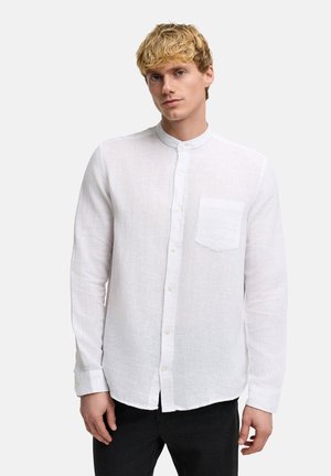 Young man with short blonde hair wearing a white button-up shirt with a mandarin collar and black pants, standing against a plain background.