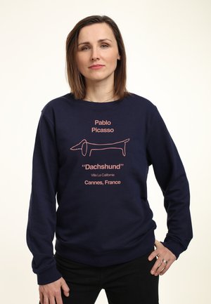 Kvinde med langt brunt hår iført en navyblå sweatshirt med en pink gravhund-tegning og tekst, der refererer til Pablo Picasso og Cannes, Frankrig.