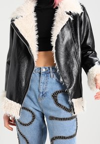 Svart faux läderjacka med mjuk krämfärgad faux pälskrage och manschetter, dragkedja framtill, kombinerad med ljusblå denimjeans med dekorativ sömnad.