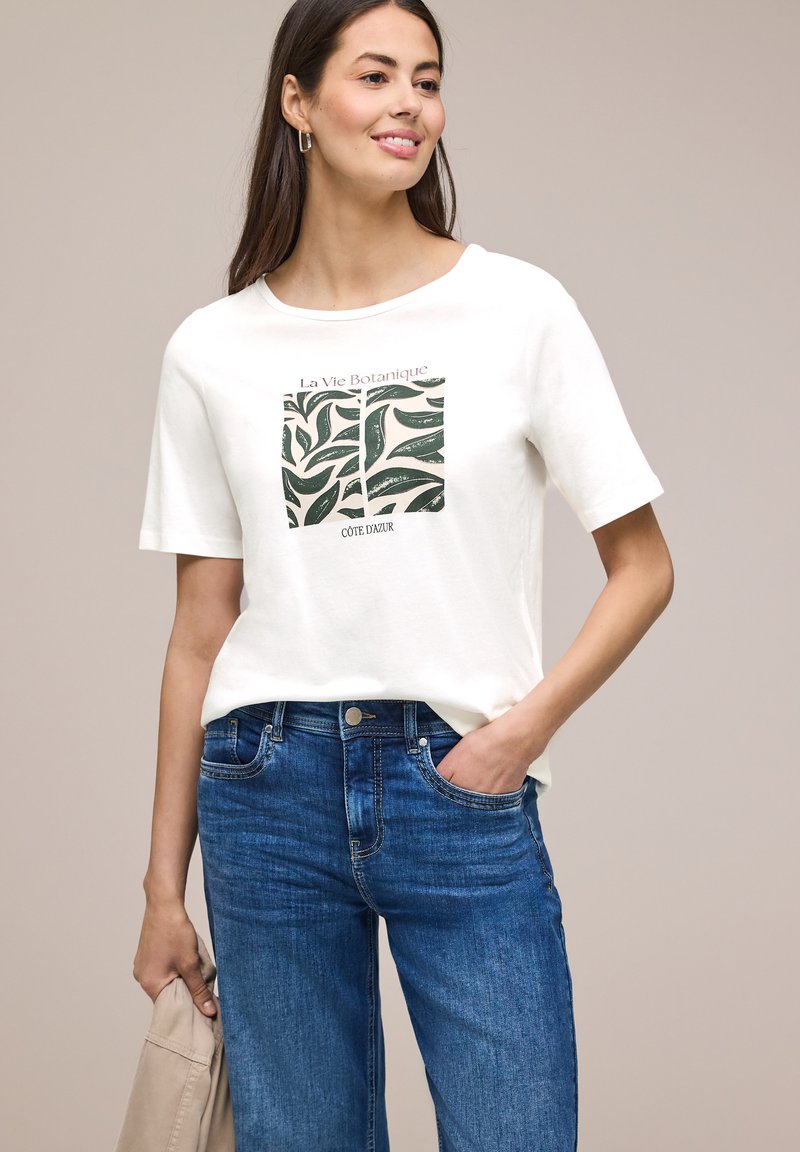 Femme souriante portant un T-shirt blanc avec un motif de feuille verte et un texte, jean bleu, tenant une veste beige sur fond neutre.