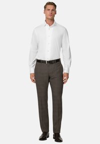 Uomo in piedi di fronte, che indossa una camicia bianca, pantaloni a quadri marroni, cintura nera e scarpe eleganti marroni scure su uno sfondo chiaro.