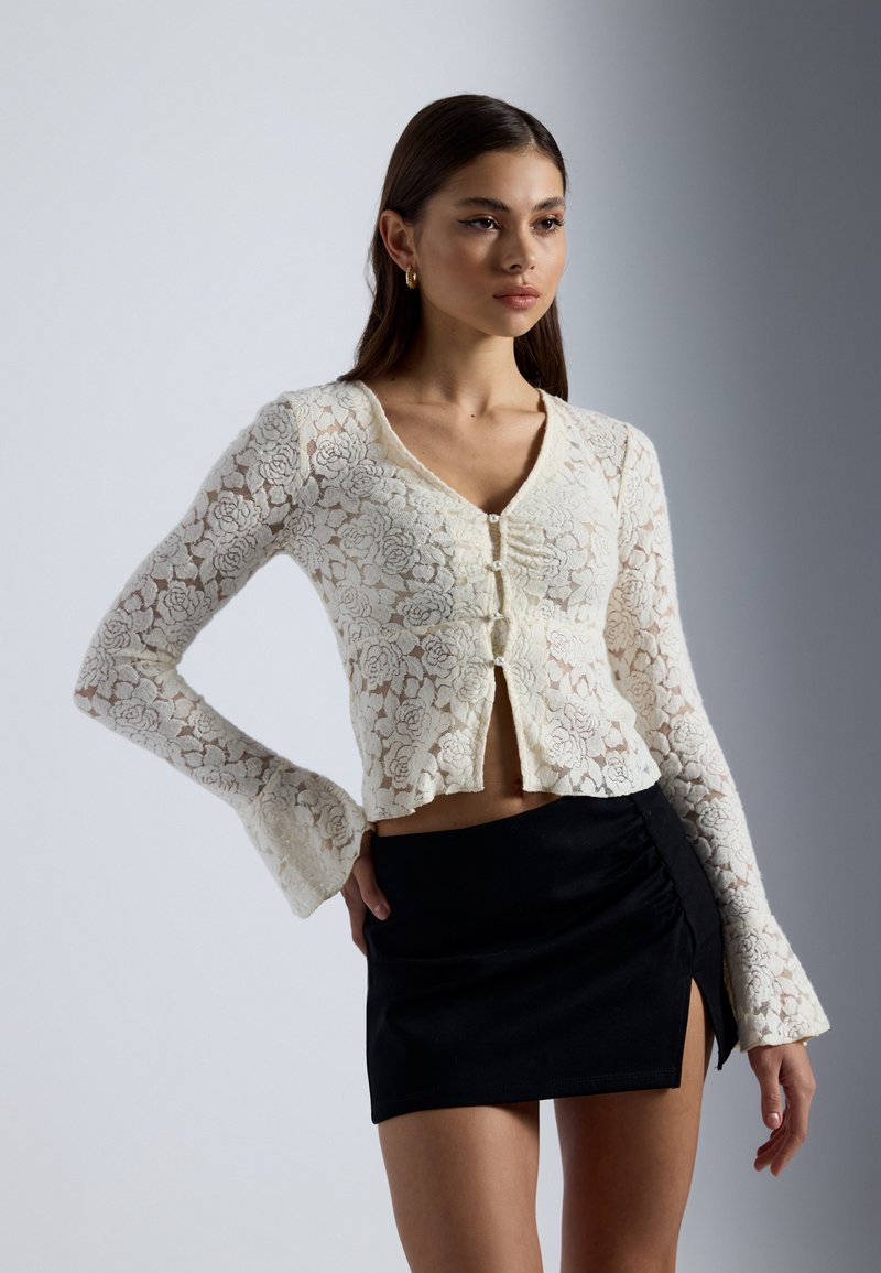 Weißer Spitzencardigan mit floralem Muster, ausgestattet mit langen Glockenärmeln und einem V-Ausschnitt, kombiniert mit einem schwarzen Minirock mit Raffung.