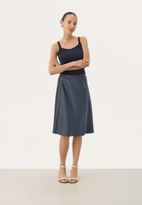 Námořnický top bez rukávů v kombinaci s lesklou tmavě šedou sukni ke kolenům, která má nařasený pas. Modelka má na sobě světlé páskové podpatky.