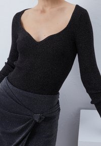 Černý žebrovaný top s dlouhými rukávy a širokým výstřihem do V, kombinovaný s tmavě šedou sukní s uzlovým detailem a jemným leskem.