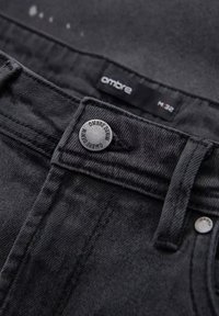 Jeans de mezclilla negros con una cinturilla visible y un botón plateado, fabricados con un tejido texturizado y una etiqueta que dice "ombre" en un diseño elegante.
