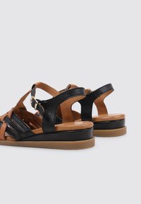 Sandalias de cuero negras y marrón con tiras cruzadas, parte trasera abierta, tacón de cuña bajo y un adorno de hebilla en la correa del tobillo. Textura suave.