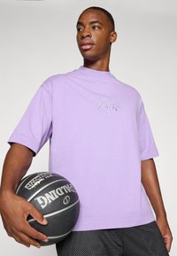 Purple short-sleeve t-shirt with "Los Angeles Lakers" embroidered logo. The model holds a black basketball in textured black shorts.