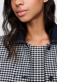 Tumesinise kraega teksatak, millel on valge ja tumesinise ruuduline muster. Sile tekstuur, esil nööpidega sulgus ja nähtavad õmblused.