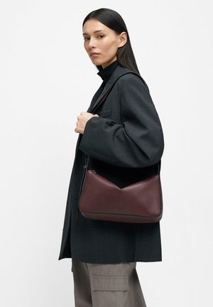 Frau mit langen dunklen Haaren trägt ein dunkles Sakko und beige Cargo-Hosen, hält eine weinrote Ledertasche über der Schulter vor einem schlichten Hintergrund.