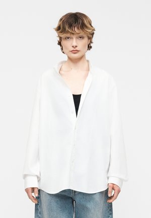 Person med kort bølget hår iført en oversized hvid skjorte over en sort top og løstsiddende blå jeans, vendt fremad med et neutralt udtryk.