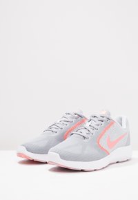 Grijze sportschoenen met een gestructureerde mesh bovenkant, roze accenten en een witte zool; ze hebben een vetersluiting en een opvallend Nike-logo.