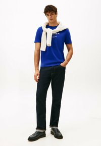 Blå T-shirt med korte ærmer og logo, parret med mørk denim jeans. En hvid kabelstrikket sweater er hængt om halsen. Sorte loafers er blevet brugt.