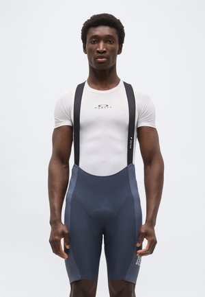 Sportos férfi fehér Oakley kompressziós pólóban és sötét kerékpáros kantáros nadrágban, egy sima háttér előtt állva.