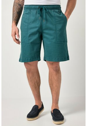 Mand iført teal snøre shorts og sorte slip-on sko står med den ene hånd i lommen og den anden arm tatoveret.