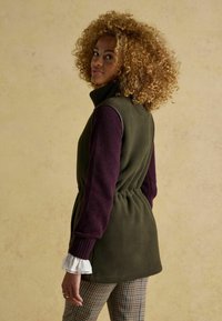 Fleecová vesta v olivovej zelenej farbe s vysokým golierom, sčítaným pásom a fialovými rukávmi. Nosí sa s kockovanými nohavicami s bielymi manžetami.