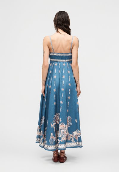 Γυναίκα με μακριά σκούρα μαλλιά που φοράει μπλε φόρεμα maxi με λεπτές ράντες, διακοσμημένο με ροζ και λευκά σχέδια, και καφέ δερμάτινα παπούτσια με κορδόνια, στέκεται μπροστά σε λευκό φόντο.