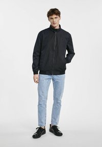 Granatowa, lekka kurtka z stojącym kołnierzem, zapięciem na zamek, elastycznymi mankietami i subtelnymi czerwonymi detalami, połączona z jasnoniebieskimi dżinsami i czarnymi butami.