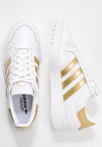 Bílé tenisky Adidas se zlatými pruhy a akcenty na podpatku, zobrazené z vrchu a z boku na bílém pozadí.