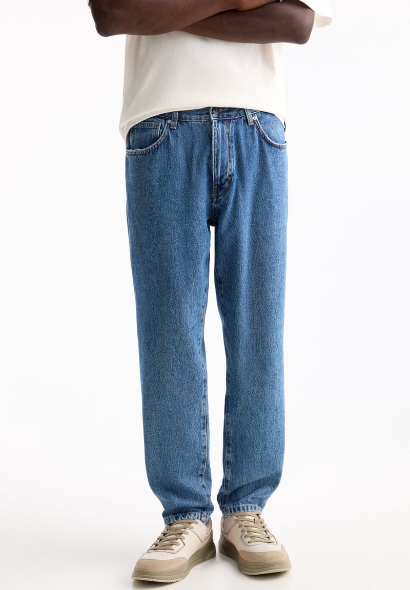 Personne portant un jean droit bleu clair, des baskets beige et un t-shirt blanc, les bras croisés, debout sur un fond blanc.