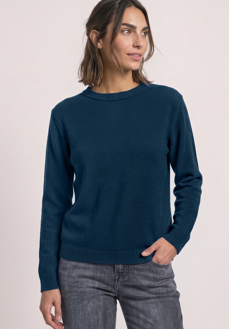 Femme portant un pull bleu foncé à manches longues et un jean gris, debout avec une main dans la poche, regardant sur le côté.