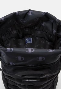 Sac à dos Champion noir ouvert montrant un compartiment principal spacieux avec une poche élastique intérieure et une doublure imprimée du logo sur toute la surface.