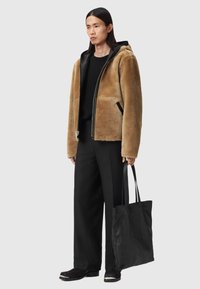 Braune Lammfelljacke mit Reißverschluss und Kapuze. Kombiniert mit einem schwarzen Pullover, lockeren schwarzen Hosen und einer strukturierten schwarzen Tragetasche.