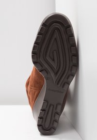 Bruna mocka ankelboots med en texturerad svart gummisula, som har ett vågigt mönster och storlek markerad som 37. Sidovyn illustrerar designen.