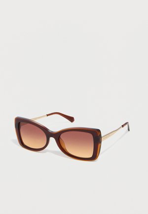 Okulary przeciwsłoneczne w kształcie kociego oka, ze szkłami w brązowym gradiencie, oprawką w kolorze żółwia i złotymi zausznikami na białym tle.