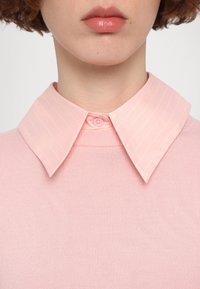 Gros plan d'une personne portant un pull côtelé rose clair superposé à une chemise rose clair à col avec des fines rayures blanches.