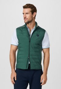 Homme portant une veste matelassée verte par-dessus un polo à manches courtes blanc et un pantalon bleu marine, regardant légèrement sur le côté.