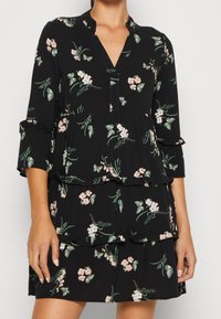 Schwarzes Blumenkleid mit V-Ausschnitt, Dreiviertelärmeln und gestuften Rüschen, verziert mit grünen Blättern und pinken Blumen. Weicher, leichter Stoff.