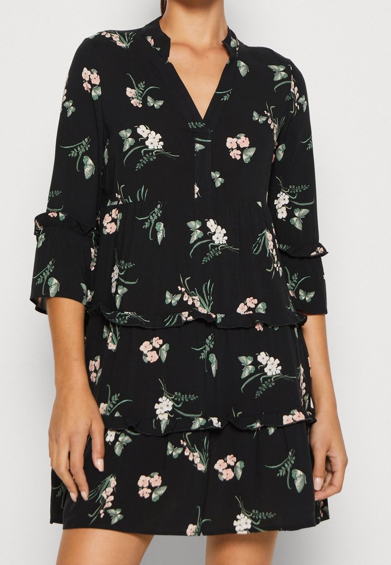 Schwarzes Blumenkleid mit V-Ausschnitt, Dreiviertelärmeln und gestuften Rüschen, verziert mit grünen Blättern und pinken Blumen. Weicher, leichter Stoff.