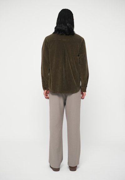 Camisa de corduroy verde-oliva com pregas nas costas, detalhes de punhos com botão, combinada com calças largas de beige claro, vista de costas.