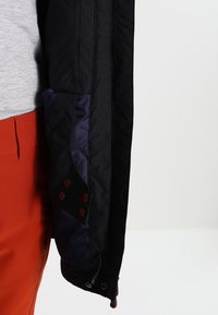 Zwarte jas met een marineblauwe zak met oogjes, ritsdetail en een gestructureerde stof. Gecombineerd met rode broek.