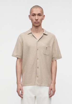 Homme à la tête rasée portant une chemise beige à manches courtes avec une poche poitrine et un pantalon blanc, debout devant un fond uni.