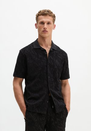 Blonde man met kort krullend haar, gekleed in een zwart kortemouwen overhemd met knopen en een bijpassende broek met patroon, handen in de zakken.