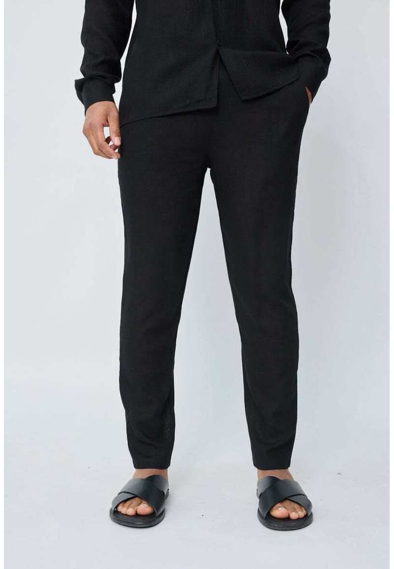 Chemise en lin noir et pantalon, texture lisse, design ajusté, manches longues, poches latérales, porté avec des sandales à glissière noires mates.