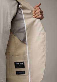 Una mano tiene aperto un giubbotto beige, mostrando la tasca interna con etichette in tessuto nero che leggono "Strellson" e "Marlane."