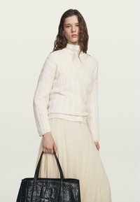 Maglione bianco lavorato a maglia con scollo a V e orlo a costine, abbinato a una gonna crema. Borsa in pelle di cocodrillo nera tenuta nella mano destra.