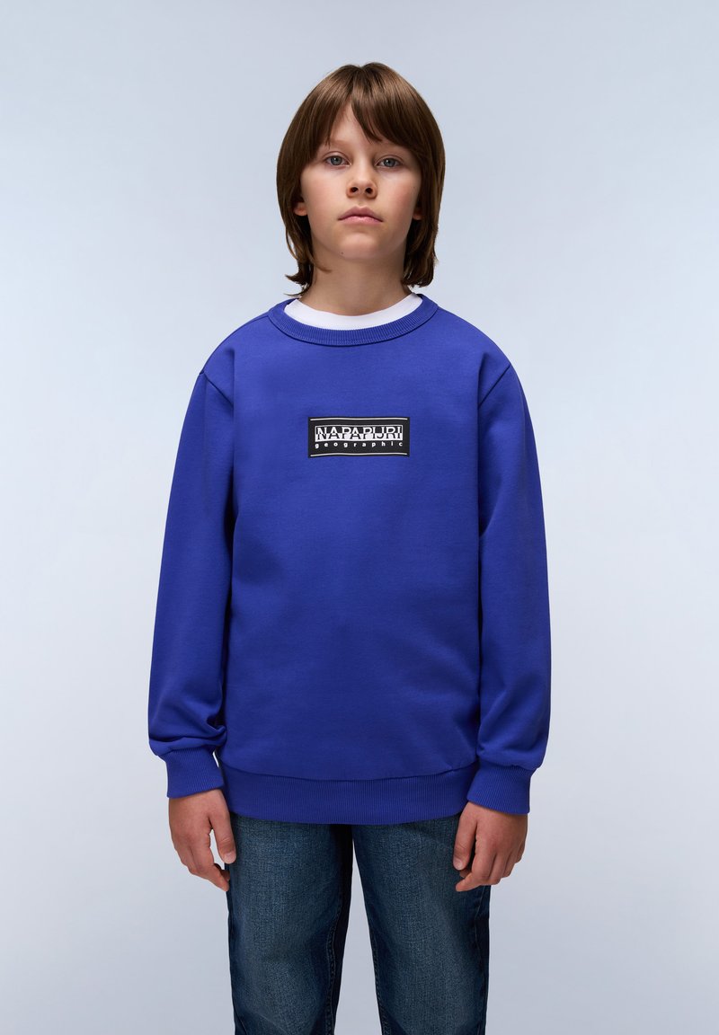Sweatshirt azul com punhos canelados, apresentando um patch de logo preto e branco no peito. Emparelhado com jeans azuis, contra um fundo claro.