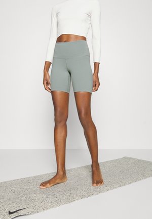 Mujer con pantalones ciclistas de cintura alta grises y una camiseta blanca de manga larga recortada, de pie descalza sobre una esterilla de yoga moteada.