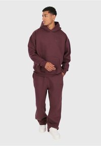 Uomo che indossa una felpa cappuccio Bordeaux scuro e pantaloni della tuta abbinati con sneakers bianche, in piedi contro uno sfondo chiaro e semplice.