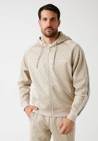 Sudadera con cremallera beige con un patrón repetido, cordones y puños acanalados. Presenta rayas contrastantes en las mangas y bolsillos laterales.